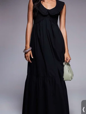 Svart långklänning med volangkrage - Svart långklänning med bred volangkrage och ärmlös design. Klänningen har en luftig passform och är tillverkad i ett mjukt tyg, perfekt för sommarens varma dagar. 