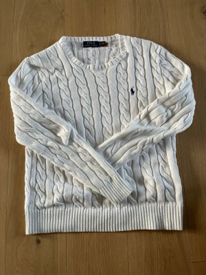 Vit kabelstickad tröja Polo Ralph Lauren - Kabelstickad vit tröja från Polo Ralph Lauren med rund halsringning och ribbade muddar. Klassisk marinblå logga broderad på bröstet. Tröjan har en tidlös design och är perfekt för lager-på-lager-stil.