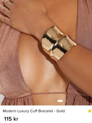 Guldigt armband - Snyggt och modernt brett cuff-armband i blankt guldfärgat metallmaterial. Aldrig använt, helt ny, kommer i orginalförpackning. 