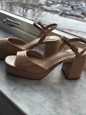 Beiga öppen tå klackar från H&M - Säljer ett par snygga beiga sandaler från H&M med breda remmar och justerbart spänne runt ankeln. Skorna har hög blockklack och platåsula i glansigt material som ger en trendig look. Perfekta för dig som vill ha både höjd och komfort.