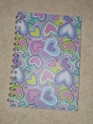 Skriv block - Säljer ett spiralblock med omslag i pastellfärger och massor av hjärtan i lila, blått, rosa och gult. En defekt finns- se bild. 