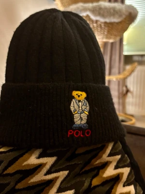 Svart ribbstickad mössa Polo Bear - Svart ribbstickad mössa från Polo Ralph Lauren med uppvikt kant. På framsidan finns den ikoniska Polo Bear-broderingen i färg, där björnen bär kostym och texten POLO i rött under. Perfekt accessoar för kalla dagar.