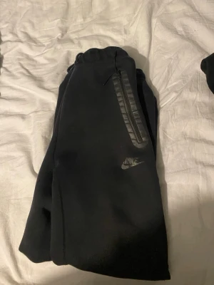 Svarta mjukisbyxor från Nike - Svarta mjukisbyxor från Nike med dragsko i midjan och två dragkedjeförsedda fickor framtill. Byxorna har en avslappnad passform och Nike-logga på vänster ben. Perfekta för chill dagar eller träning.