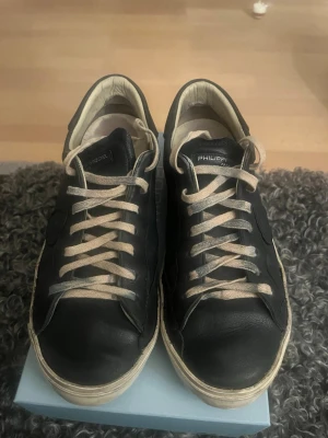 Svarta Philippe Model sneakers - Snygga svarta sneakers från Philippe Model med beige snörning och vit sula. Skorna har en klassisk låg profil och är tillverkade i läder med detaljer i samma färg. Perfekta för dig som gillar stilrena och tidlösa sneakers.