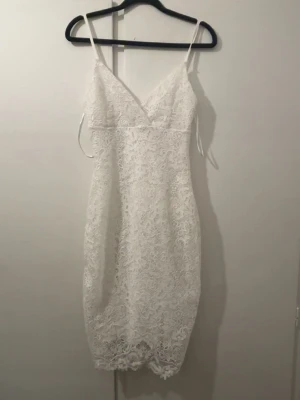 Vit spetsklänning med smala axelband - Säljer en elegant vit spetsklänning med smala axelband och v-ringning. Klänningen är figurnära och har ett vackert spetsmönster över hela plagget. Perfekt för sommarens alla tillfällen och studentfirande.