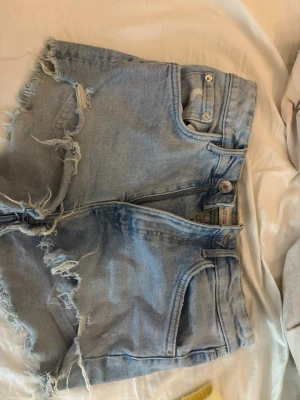 Ljusblå denimshorts med fransar - Säljer ett par ljusblå denimshorts med råa, fransiga kanter och slitningar. Klassisk femficksmodell med dragkedja och knapp framtill. Perfekta för sommaren och ger en avslappnad vibe.