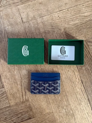 Blå Goyard Plånbok - Snyggt blått kortfodral från Goyard med klassiskt mönster i vitt och grått. Tillverkad i canvas med läderdetaljer. Kommer med original grön ask och Goyard-kort. Perfekt för kort och ID.