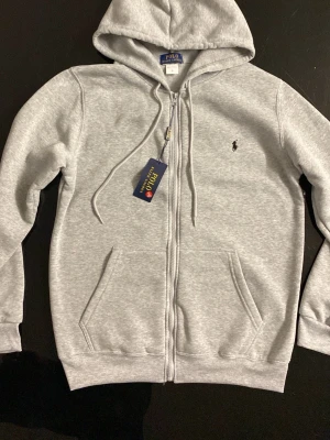 Ralph Lauren zip hoodie - Säljer en grå zip-hoodie från Ralph Lauren i mycket bra skick. Perfekt för både vardag och fritid klassisk design som aldrig går ur stil!