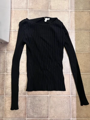 Svart ribbstickad långärmad tröja - En svart ribbstickad tröja med lång ärm och rund halsringning. Tröjan är figurnära och har ett klassiskt, stilrent utseende som passar perfekt till jeans eller kjol. Materialet är mjukt och stretchigt, vilket gör den bekväm att bära hela dagen.