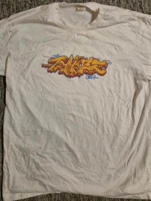 Vit t-shirt med graffiti-tryck XL/xxl - Säljer en vit t-shirt med ett stort, färgstarkt graffiti-tryck i gult, orange och blått på bröstet. T-shirten har rund hals och korta ärmar. Perfekt för dig som gillar streetwear och unika prints. Storleken är XL/xxl