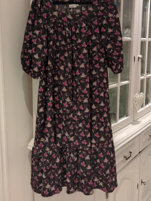 Blommig kortklänning från Isolde - Söt klänning från Isolde med svart botten och blommigt mönster i rosa och beige. Klänningen har puffiga korta ärmar och en lös, flowy passform. Volang nedtill. Perfekt för dig som gillar romantiska vibes och vill ha något bekvämt men ändå stylish.