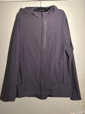 Grå vindjacka från Under Armour - Snygg grå vindjacka från Under Armour med huva och dragkedja framtill. Jackan har logga och text på bröstet, två sidofickor och är tillverkad i ett lätt, tekniskt material som passar perfekt för aktiva dagar utomhus.