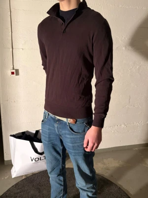 Mörkbrun långärmad pikétröja - Säljer en mörkbrun långärmad pikétröja med krage och två knappar vid halsen. Tröjan har ribbade muddar vid ärmslut och nederkant. Perfekt för dig som gillar en stilren och enkel look.