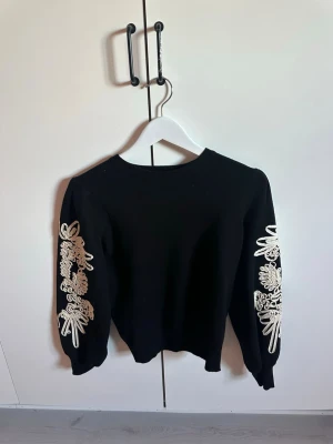 Svart stickad tröja med brodyr - Snygg svart stickad tröja med vita broderade blommor längs båda ärmarna. Tröjan har rund halsringning och långa ärmar med ribbade muddar. Perfekt för dig som vill ha något unikt och trendigt i garderoben.
