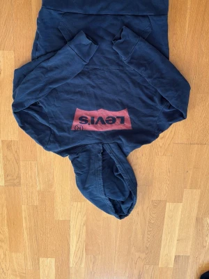 Mörkblå hoodie från Levi's - Snygg mörkblå hoodie från Levi's med stor röd logga på bröstet. Klassisk modell med huva och känguruficka framtill. Perfekt för en avslappnad och trendig stil. Tillverkad i mjukt bomullsmaterial.