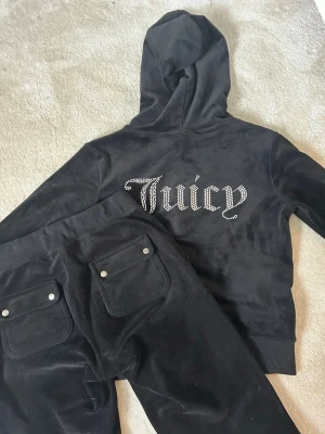 Svart Juicy Couture velourset - Säljer ett svart velourset från Juicy Couture med hoodie och byxor. Hoodien har huva och stora strasstenar som bildar texten 'Juicy' på ryggen. Byxorna har fickor med knappar bak. Perfekt för chill dagar hemma eller när du vill vara extra comfy.                    Tröjan är storlek S & byxorna är storlek XS