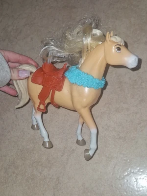 Leksakshäst - Säljer en leksakshäst i plast med ljusbrun kropp, vit man och svans samt vita ben. Hästen har en rödbrun sadel och ett blått stickat halsband. Perfekt för dig som gillar hästar och vill ha en snygg detalj till samlingen.