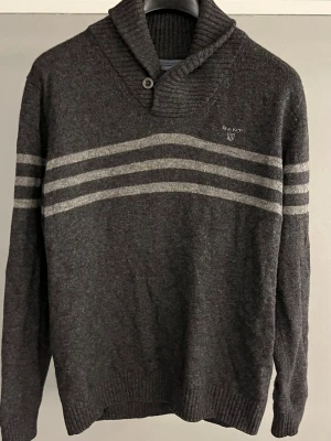 Mörkgrå stickad tröja från GANT L-M - Snygg stickad tröja från GANT med bred ribbad sjalkrage och knappdetalj. Tröjan har tre ljusgrå ränder över bröstet och armarna samt broderad logga på bröstet. Den är i storlek L men sitter snarare som en M nästa till och med S. Hör gärna av dig om du undrar något eller vill se fler bilder.🤩☀️priset kan diskuteras.