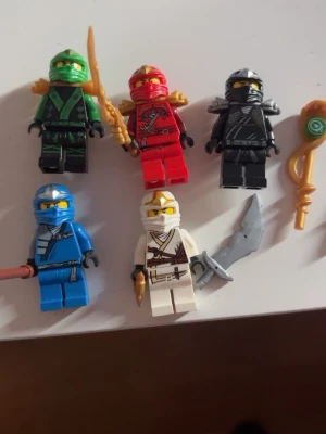 LEGO Ninjago figurer med vapen - Fem LEGO Ninjago minifigurer i färgerna grön, röd, svart, blå och vit. Figurerna har detaljerade dräkter och tillhörande vapen som svärd och stav. Perfekt för dig som gillar att bygga och leka med Ninjago-karaktärer.