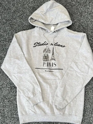 Grå hoodie Studio Silvano Paris - Snygg grå hoodie från Studio Silvano med tryck av Eiffeltornet och texten 'Paris' på bröstet. Hoodien har huva med dragsko och en stor magficka. Tillverkad i mjukt sweatshirtmaterial, perfekt för en avslappnad streetstyle-look.