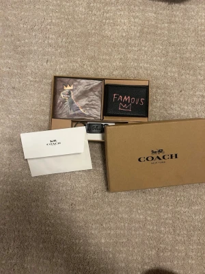Coach x Basquiat svart plånbok - Snygg svart plånbok från Coach i samarbete med Basquiat. Plånboken har coolt tryck med texten 'FAMOUS' och en ikonisk krona på framsidan. Tillverkad i äkta läder med flera kortfack och stilren Coach New York-logga inuti. Perfekt för dig som gillar street art och exklusiva detaljer.