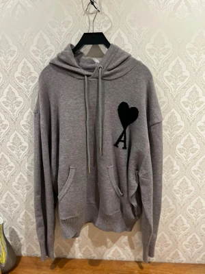 Grå Ami Paris hoodie - Snygg grå hoodie från Ami Paris med stor svart hjärtlogga och bokstaven A på bröstet. Hoodien har huva med dragsko, känguruficka och ribbade muddar. Tillverkad i mjukt stickat material som känns riktigt skönt mot huden.