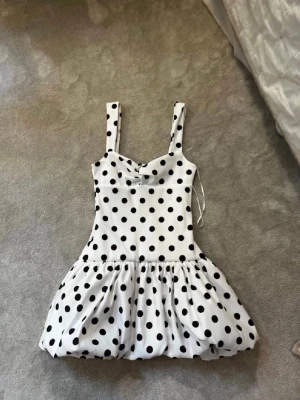 Vit prickig kortklänning - Söt vit kortklänning med svarta prickar och breda axelband. Klänningen har en puffig kjol och markerad midja som ger en lekfull siluett. Perfekt för dig som vill sticka ut med ett klassiskt mönster och trendig form.