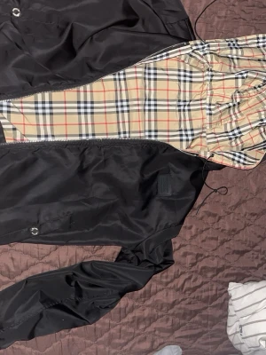 Svart vindjacka från Burberry - Snygg svart vindjacka från Burberry med klassiskt beige-rutigt foder och huva. Jackan har dragkedja framtill och justerbar huva med snörning. Perfekt för dig som vill ha en stilren och ikonisk look.