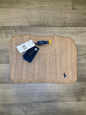 Beige kabelstickad tröja Polo Ralph Lauren - Snygg beige kabelstickad tröja från Polo Ralph Lauren med rund halsringning och klassisk marinblå logga broderad på bröstet. Tröjan har långa ärmar och ribbade muddar vid ärmslut och nederkant. Perfekt för en stilren och avslappnad look.
