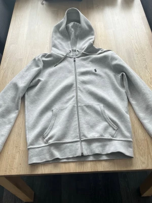 Ljusgrå zip hoodie från Polo Ralph Lauren - Snygg ljusgrå hoodie med dragkedja från Polo Ralph Lauren. Klassisk design med huva, två fickor fram och den ikoniska lilla broderade loggan på bröstet. Tillverkad i mjuk bomullsblandning som känns skön mot huden. Perfekt för en avslappnad och stilren look.