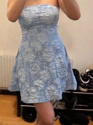 Ljusblå axelbandslös klänning - Söt axelbandslös klänning i ljusblått med blommigt mönster och lätt utsvängd kjol. Perfekt för sommarens alla tillfällen och ger en fräsch och ungdomlig look.