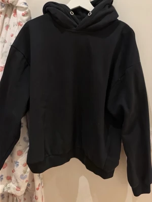 Svart basic hoodie med huva - En enkel svart hoodie med huva och dragsko. Hoodien har en avslappnad passform, ribbade muddar vid ärmar och nederkant samt en klassisk look utan tryck eller logga. Perfekt för en chill och stilren vardagsoutfit.