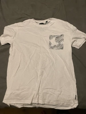 Vit t-shirt med mönstrad ficka - Säljer en vit t-shirt med korta ärmar och en snygg bröstficka med svartvitt palmbladsmönster. T-shirten har en klassisk passform och är gjord i mjuk bomull, perfekt för en avslappnad stil.