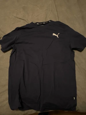 Mörkblå Puma t-shirt - Snygg mörkblå t-shirt från Puma med klassisk vit logga på bröstet. T-shirten har rund hals och korta ärmar, perfekt för en avslappnad och sportig stil. 