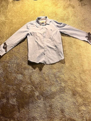 Ljusblå skjorta med knappar - Klassisk ljusblå skjorta med krage och knäppning framtill. Skjortan har långa ärmar och är tillverkad i ett tunt bomullstyg. Perfekt för en clean och stilren look.