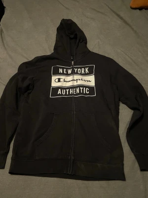 Svart hoodie från Champion - Svart hoodie med dragkedja från Champion. Tryck på bröstet med texten 'New York Champion Authentic' i vitt. Klassisk huva och två fickor framtill. Perfekt för en avslappnad streetwear-look.