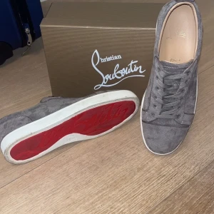 Gråa sneakers från Christian Louboutin - Snygga gråa sneakers i mocka från Christian Louboutin med den ikoniska röda sulan. Skorna har klassisk låg design, rund tå och vita kanter på sulan. Perfekta för dig som vill ha något exklusivt och stilrent i garderoben. Använda 5 gånger