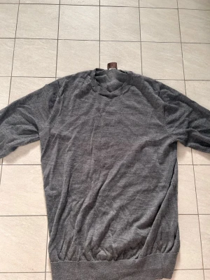 Grå stickad tröja med rund hals - En enkel och stilren grå stickad tröja med rund halsringning och långa ärmar från Singular Society. Tröjan har ribbstickade muddar vid ärmslut och nederkant, vilket ger en klassisk look. Perfekt att matcha med jeans eller kjol för en avslappnad stil.