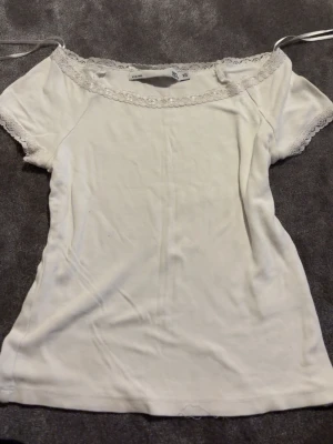 Vit topp med spetsdetaljer från lager 157 - Söt vit topp från Lager 157 med korta ärmar och spetsdetaljer längs halsringningen och ärmsluten. Toppen har en offshoulder-design med tunna band som knyts på axlarna. Perfekt för en fräsch och feminin look.