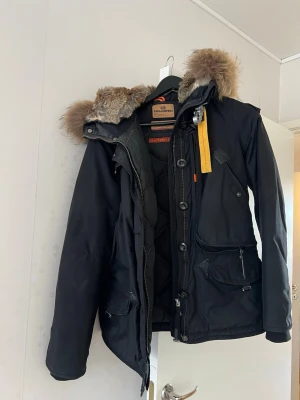 Svart dunjacka Parajumpers pälsluva - Säljer en svart vinterjacka från Parajumpers med avtagbar huva och pälsdetalj. Jackan har flera fickor, gul rem vid kragen och patch på axeln. Materialet är slitstarkt och jackan är vadderad för extra värme. Perfekt för kalla dagar. Strl M 