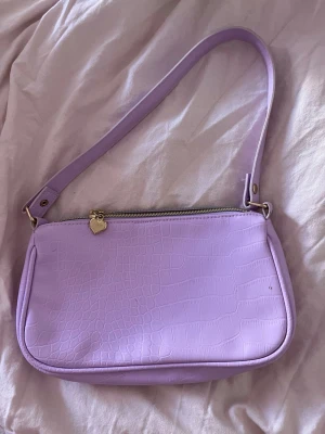 Ljuslila axelväska med hjärtdetalj - Säljer en ljuslila axelväska i fejkat krokodilskinn med en söt hjärtformad dragkedjeberlock 💕 Perfekt för dig som vill ha en trendig och färgglad accessoar, väldigt vårig 🌸Priset går att diskutera 🥰
