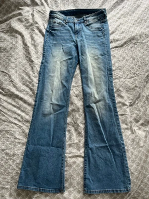Blå bootcut jeans med slitningar - Säljer ett par ljusblå bootcut jeans med snygga slitningar framtill och bakfickor. Jeansen har klassisk femficksmodell och normal midja. Perfekta för dig som gillar retrovibbar och vill ha ett par jeans med lite vidare ben. Materialet är mjukt denim.