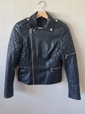 Svart skinnjacka från Guess - Snygg svart skinnjacka från Guess med quiltade detaljer på ärmar och axlar. Jackan har snedställd dragkedja, två fickor med dragkedja och axelklaffar med tryckknappar. Klassisk bikerstil som ger en cool look.