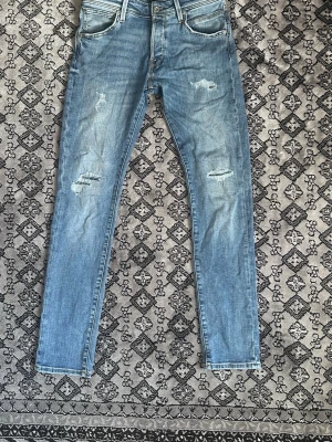 Ljusblå slitna jeans från Jack and Jones  - Säljer ett par ljusblå skinny jeans med slitningar på låren och knäna. Klassisk femficksmodell med normalhög midja och smal passform. Jeansen har en brun patch bak i midjan och är tillverkade i stretchigt denimtyg för extra komfort. Dessa byxor är använda vardagligen tidigare men inga syn på skavanker eller liknade.