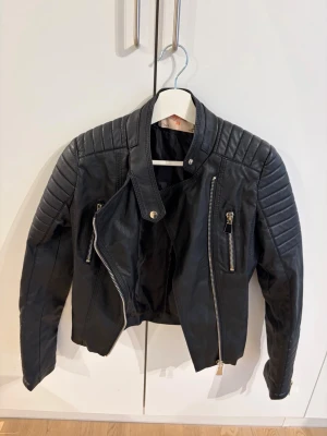 Svart skinnjacka från Voyelles - Snygg svart skinnjacka från Voyelles med coola detaljer som quiltade axlar, snedställd dragkedja och två dragkedjefickor framtill. Jackan har en hög krage med tryckknapp och en figurnära passform som ger en edgy look.