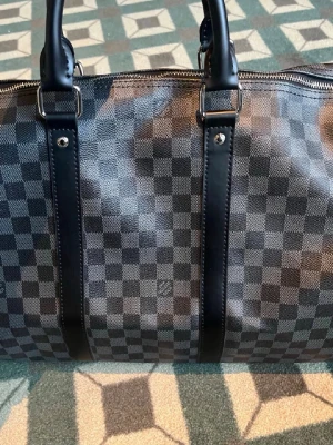 Louis Vuitton Damier Keepall väska - Snygg resväska från Louis Vuitton i klassiskt Damier-mönster i svart och grått. Väskan har dubbla handtag, silverfärgade metalldetaljer och dragkedja med LV-logga. Tillverkad i slitstarkt canvasmaterial med läderdetaljer. Perfekt för weekendresor.