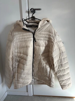 Beige dunjacka med huva - Snygg beige dunjacka med huva och dragkedja framtill. Jackan har quiltad design och två sidofickor. Perfekt för kalla vinterdagar och ger en stilren look. Passar dig som vill ha en lätt och varm jacka.
