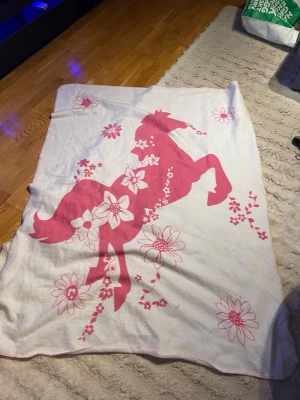 Vit filt med rosa häst och blommor - Mysig vit filt med stort rosa hästmotiv och blommor. Filtens form är rektangulär och den har dekorativa sömmar längs kanterna. Materialet känns mjukt, troligen fleece eller bomullsmix. Perfekt för att mysa i soffan eller sängen.