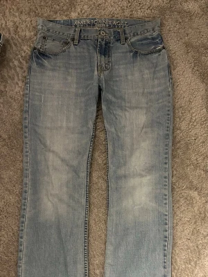 Ljusblå jeans från American Eagle - Säljer ett par ljusblå jeans från American Eagle med raka ben. Jeansen har slitna detaljer och kontrastsömmar, samt motiv på bakfickor. Storlek 32/32