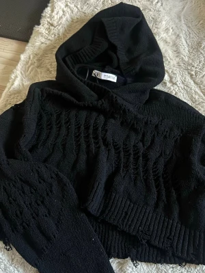 Svart stickad hoodie från Zara - Säljer en svart stickad hoodie från Zara i storlek XS. Hoodien har en croppad passform, ribbade muddar och ett coolt hålmönster över hela plagget. 